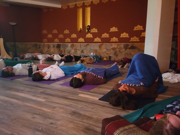 Cozy restorative yoga class with participants lying on mats and wrapped in blankets for guided relaxation in a dim, patterned-walled studio with wood floors.