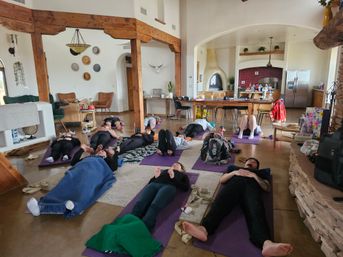 Group relaxation session on yoga mats in a sunny open-plan living room with exposed wooden beams, high ceilings, kiva-style fireplace and kitchen