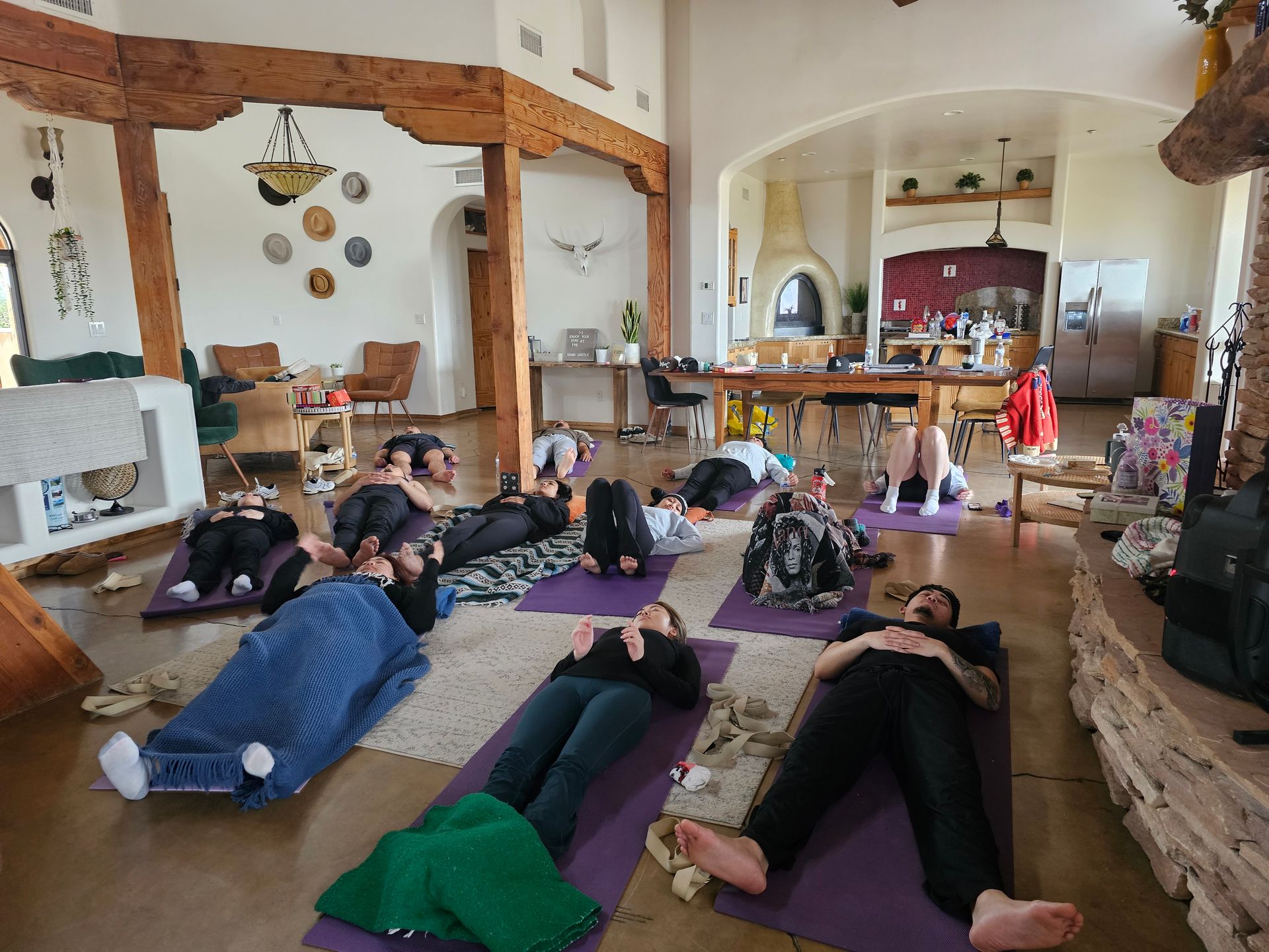 Group relaxation session on yoga mats in a sunny open-plan living room with exposed wooden beams, high ceilings, kiva-style fireplace and kitchen