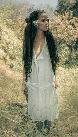 Boho outdoor portrait of a woman with long dreadlocks in a white sundress, standing in a sunlit meadow with a flower tucked behind her ear, looking to the side.