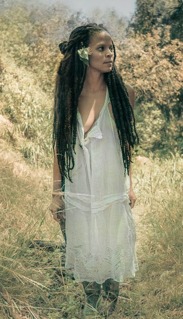 Boho outdoor portrait of a woman with long dreadlocks in a white sundress, standing in a sunlit meadow with a flower tucked behind her ear, looking to the side.