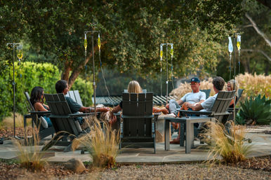 Adults relaxing on Adirondack chairs around a fire pit on a shaded outdoor garden patio, each with an IV drip bag on a stand for an outdoor wellness session.