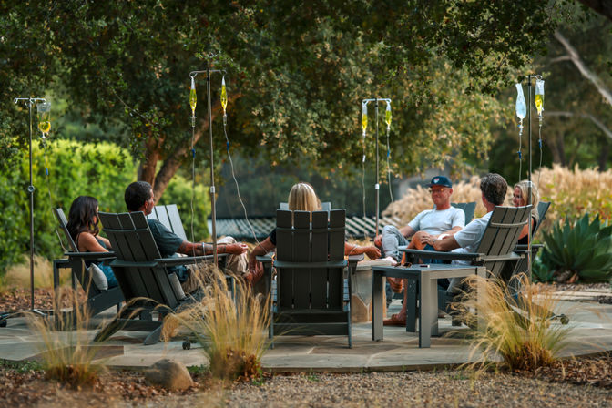 Adults relaxing on Adirondack chairs around a fire pit on a shaded outdoor garden patio, each with an IV drip bag on a stand for an outdoor wellness session.