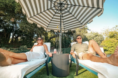 Two adults relaxing on white-cushioned lounge chairs under a large striped patio umbrella in a sunny garden, drink on a small side table between them.