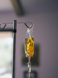 Close-up of a golden IV infusion bag hanging from a stainless-steel pole, drip chamber and tubing visible against a softly blurred clinical background.