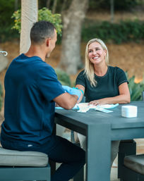 Smiling woman receiving an outdoor blood draw from a gloved healthcare worker at a patio table