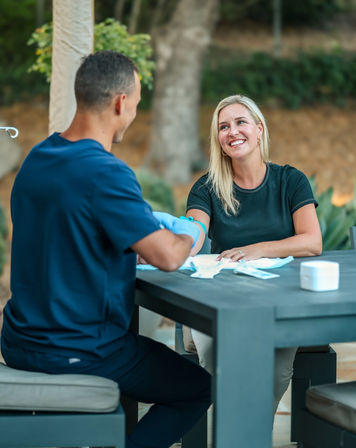 Smiling woman receiving an outdoor blood draw from a gloved healthcare worker at a patio table
