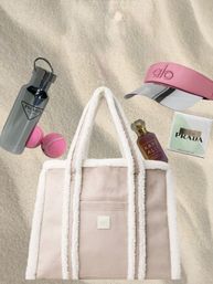Beach-ready plush shearling-trimmed pink tote on sand surrounded by a mirrored water bottle, pink visor, two pink tennis balls, a perfume bottle and a skincare sachet