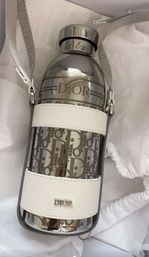 Sleek reflective designer water bottle with repeating monogram pattern, metallic cap and white leather strap carrier, photographed on white fabric