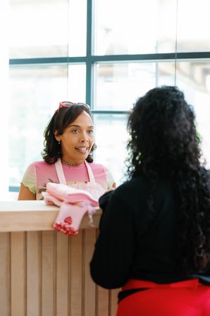 Boutique checkout scene: staff in a pink apron and sunglasses-on-head talks to a customer across a wooden counter with folded pink gloves and garments, sunlit window in the background.