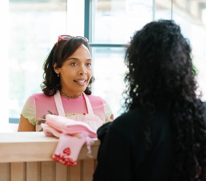 Boutique checkout scene: staff in a pink apron and sunglasses-on-head talks to a customer across a wooden counter with folded pink gloves and garments, sunlit window in the background.