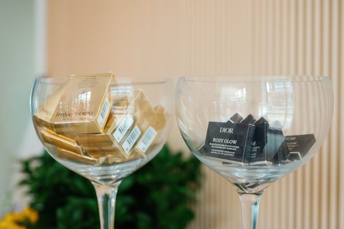 Two oversized stemmed glass bowls holding luxury makeup sample boxes — stacked gold YSL packages on the left and small black Dior "Rosy Glow" boxes on the right, displayed like candy in a chic display.
