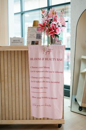 Cheerful pink floral bouquet and framed beauty samples on a light-wood salon reception counter with a pink ‘bloom & beauty bar’ banner