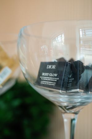 Close-up of small black cosmetic sample boxes in a large stemmed glass bowl on a boutique counter, soft window reflection and blurred greenery in the background