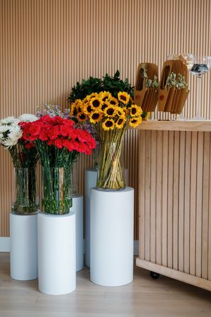 Modern floral display with sunny sunflowers, red carnations and white chrysanthemums in glass vases on white cylindrical pedestals against a vertical wood‑slat wall — stylish flower shop interior.
