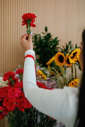 Hand holding a bright red carnation aloft above a mixed bouquet of sunflowers and carnations in an indoor flower shop display.