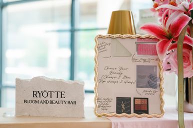 Tabletop beauty display in a bright indoor boutique featuring a textured stone welcome sign, ornate gold-framed makeup menu, and pink lilies