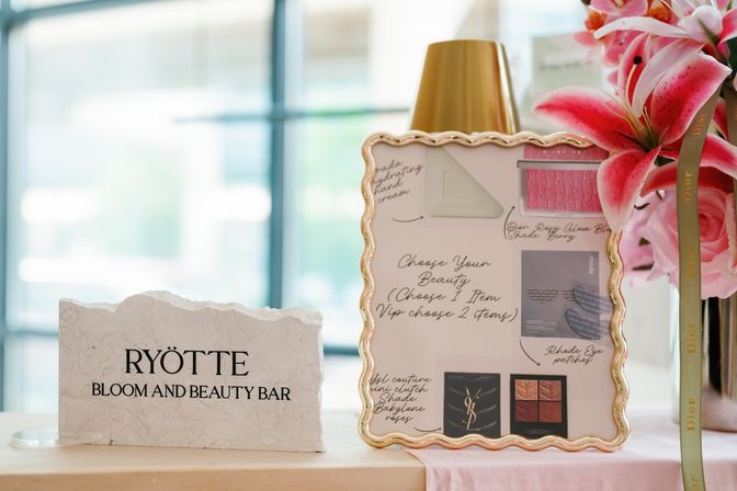 Tabletop beauty display in a bright indoor boutique featuring a textured stone welcome sign, ornate gold-framed makeup menu, and pink lilies