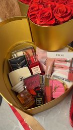 Gold heart-shaped gift box overflowing with red roses and designer beauty items — mini perfumes, lip glosses, compacts and accessories arranged as a romantic gift