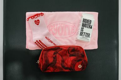 Pink workout towel on a black exercise mat with white pilates socks featuring red heart grips and ruffle trim, a small boxed water carton, and a red patterned zip pouch.