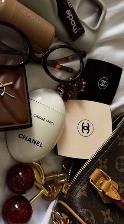 Luxury travel essentials flat lay on white fabric: tortoiseshell round glasses, heart-shaped hand cream, compact with interlocking logo, brown leather cardholder, glittery red key charms, tan monogram zip pouch and makeup tubes.