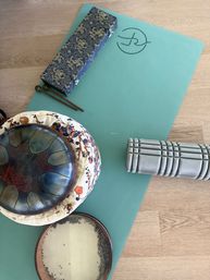 Mint yoga mat on wood floor with sound-healing instruments: steel tongue drum on a patterned cushion, frame drum, mallets in a decorative box and a gray foam roller — home meditation setup.