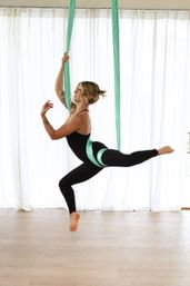 Person in black activewear suspended on mint-green aerial silk in a bright studio, holding a graceful aerial yoga pose with one leg extended