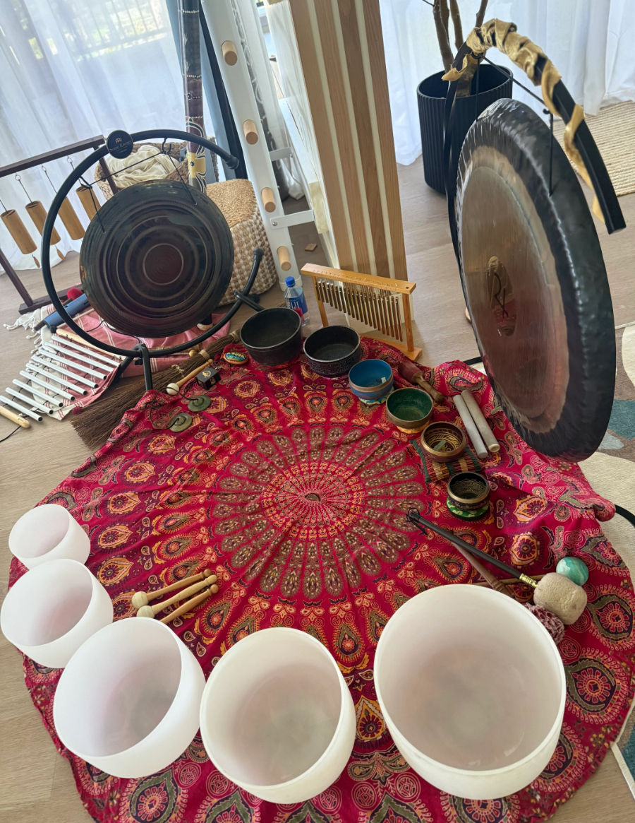 Sunlit wellness studio sound-healing setup on a red mandala cloth with crystal singing bowls, Tibetan metal bowls, two gongs, chimes and mallets in a bright indoor meditation space