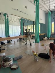 Bright, airy aerial yoga studio interior with green silk hammocks hanging from the ceiling, a mirrored wall with barre, students on mats on a wood floor and scattered wellness props and baskets.