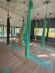 Bright aerial yoga studio with teal and gray silk hammocks hanging from the ceiling over matching teal mats on a light wood floor, mirrored wall and large windows letting in sunlight.