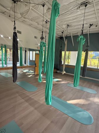 Bright aerial yoga studio with teal and gray silk hammocks hanging from the ceiling over matching teal mats on a light wood floor, mirrored wall and large windows letting in sunlight.
