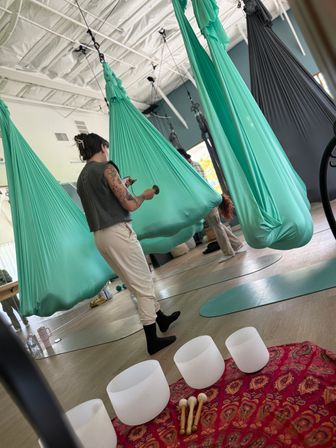 Aerial yoga studio with teal silk hammocks hanging from a high white ceiling, participants preparing on mats, and white crystal singing bowls with mallets on a patterned cloth in the foreground.