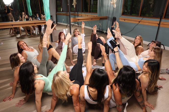 Group of women in activewear seated in a mirrored fitness studio, playfully raising legs in a synchronized circle with wooden floors, windows and a few wearing heels.