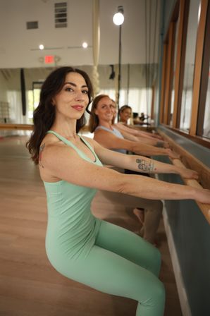 Close-up of a woman in mint-green activewear doing a barre squat with hands on a wooden barre in a bright fitness studio, two classmates lined up and smiling behind her.