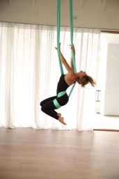 Woman practicing aerial yoga on teal silks, leaning into a playful backbend mid-air in a bright studio with sheer white curtains and light wood floor.