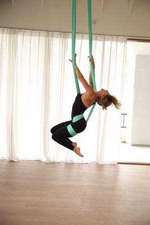 Woman practicing aerial yoga on teal silks, leaning into a playful backbend mid-air in a bright studio with sheer white curtains and light wood floor.