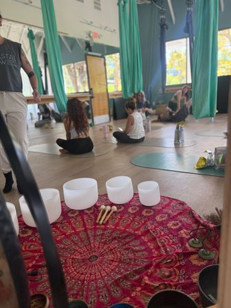 Aerial yoga studio interior with teal silk hammocks hanging from the ceiling, students seated on yoga mats, and white crystal singing bowls with mallets arranged on a red mandala cloth for a sound bath.