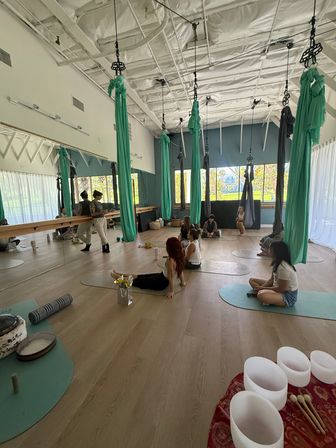 Sunlit aerial yoga studio with teal silk hammocks suspended from the ceiling, people seated on mats, mirrored wall, wooden floor and crystal singing bowls in the foreground