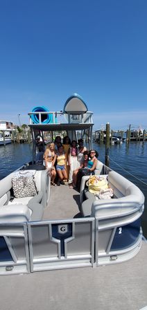 Group of people on a pontoon boat at a sunny marina — cushioned seating and bags onboard, stacked blue kayaks on the roof, wooden docks and boats in the harbor under a clear blue sky.