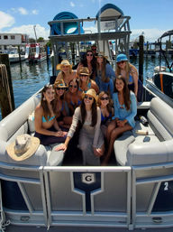 Group of women in swimsuits and sun hats posing on a pontoon boat at a sunny marina with docked boats and blue sky.