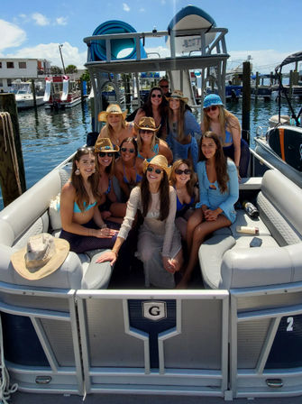 Group of women in swimsuits and sun hats posing on a pontoon boat at a sunny marina with docked boats and blue sky.