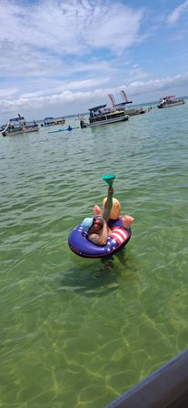 Person lounging on a stars-and-stripes inflatable tube in clear green coastal water, holding a funnel to a novelty inflatable doll; pontoon boats and swimmers in the background under a blue, partly cloudy sky — lively summer beach and boating scene.