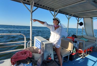 Person in a captain's hat, sunglasses, veil and lei wearing a white cover-up, striking a captain pose and pointing toward the ocean horizon from the shaded deck of a leisure boat on a sunny day.