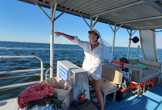Person in a captain's hat, sunglasses, veil and lei wearing a white cover-up, striking a captain pose and pointing toward the ocean horizon from the shaded deck of a leisure boat on a sunny day.