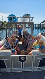 Group of friends on a pontoon boat at a sunny marina, wearing cowboy hats and leis with paddleboards stacked on the roof and waterfront docks under a blue sky.