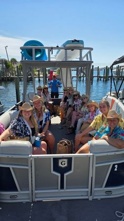 Group of friends on a pontoon boat at a sunny marina, wearing cowboy hats and leis with paddleboards stacked on the roof and waterfront docks under a blue sky.