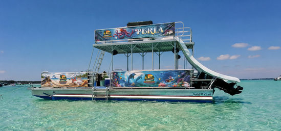 Double-decker pontoon boat with a curved water slide and colorful ocean-themed murals floating in shallow turquoise tropical water under a clear blue sky