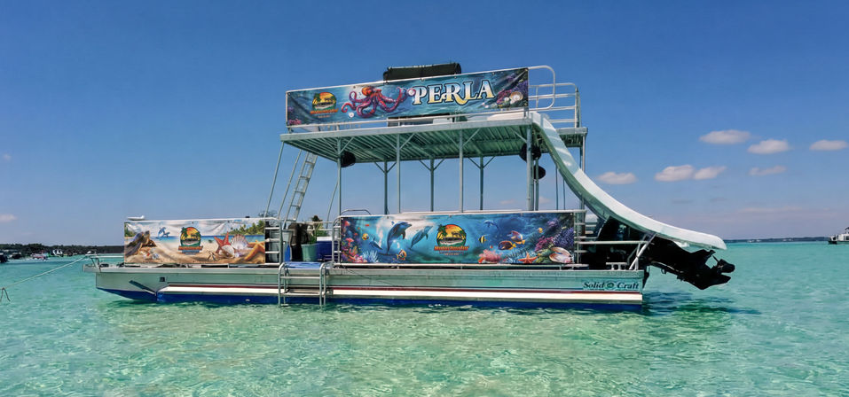 Double-decker pontoon boat with a curved water slide and colorful ocean-themed murals floating in shallow turquoise tropical water under a clear blue sky