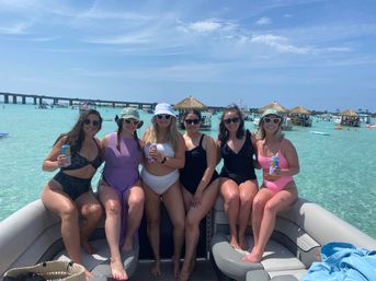 Six friends in colorful swimsuits sitting on a pontoon boat, holding drinks in shallow turquoise water near tiki huts and a bridge on a sunny blue day