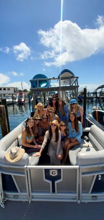 Group of smiling women in swimwear and sun hats aboard a pontoon boat at a sunny marina, blue sky and puffy clouds overhead.